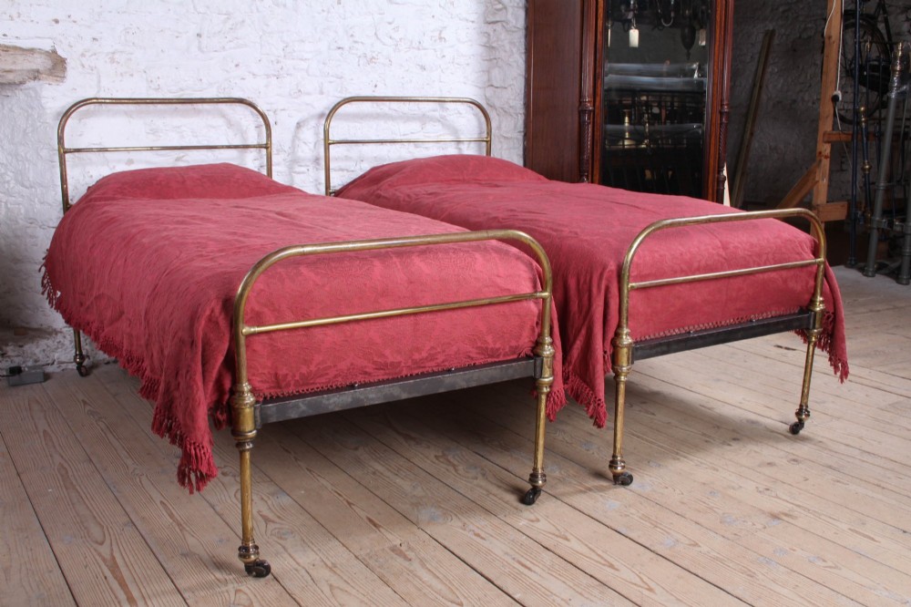 A pair of antique tubular brass single bed frames featuring rounded headboards, simple footboards, turned leg details, and brass castors.
