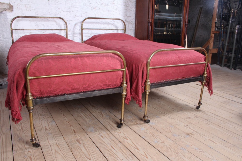 A pair of antique tubular brass single bed frames featuring rounded headboards, simple footboards, turned leg details, and brass castors. View VI