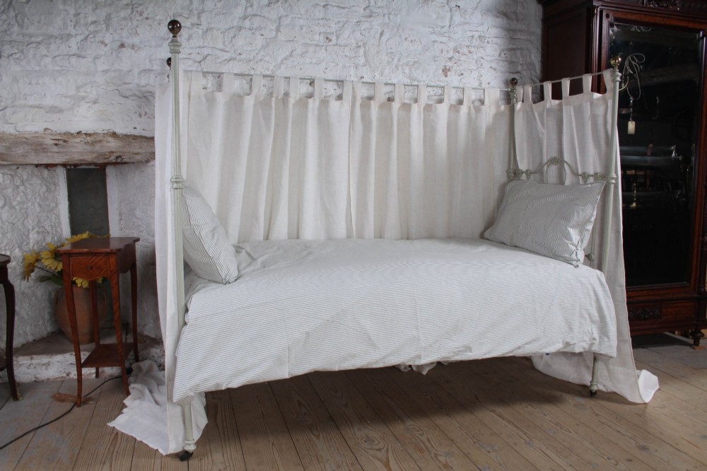 A painted single iron four-poster bed frame featuring decorative central scrollwork, brass top finials, and lightweight drapery. View IV