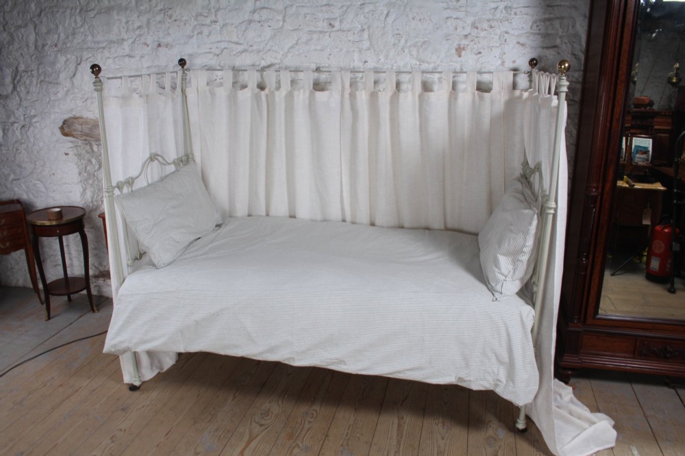 A painted single iron four-poster bed frame featuring decorative central scrollwork, brass top finials, and lightweight drapery. View V