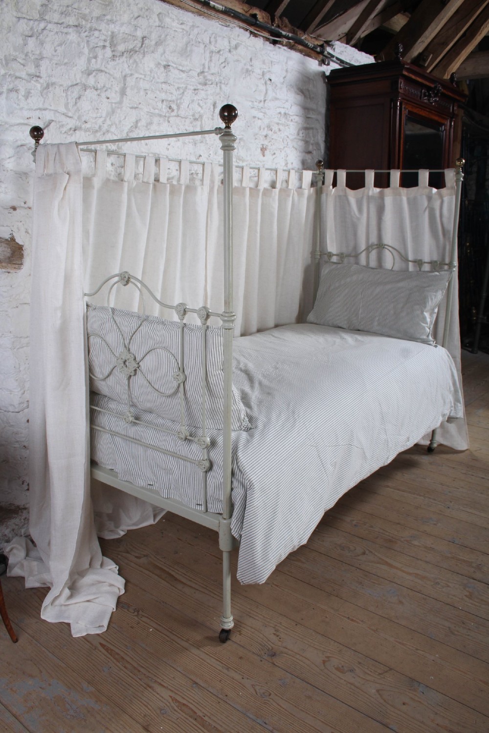 A painted single iron four-poster bed frame featuring decorative central scrollwork, brass top finials, and lightweight drapery. View VI