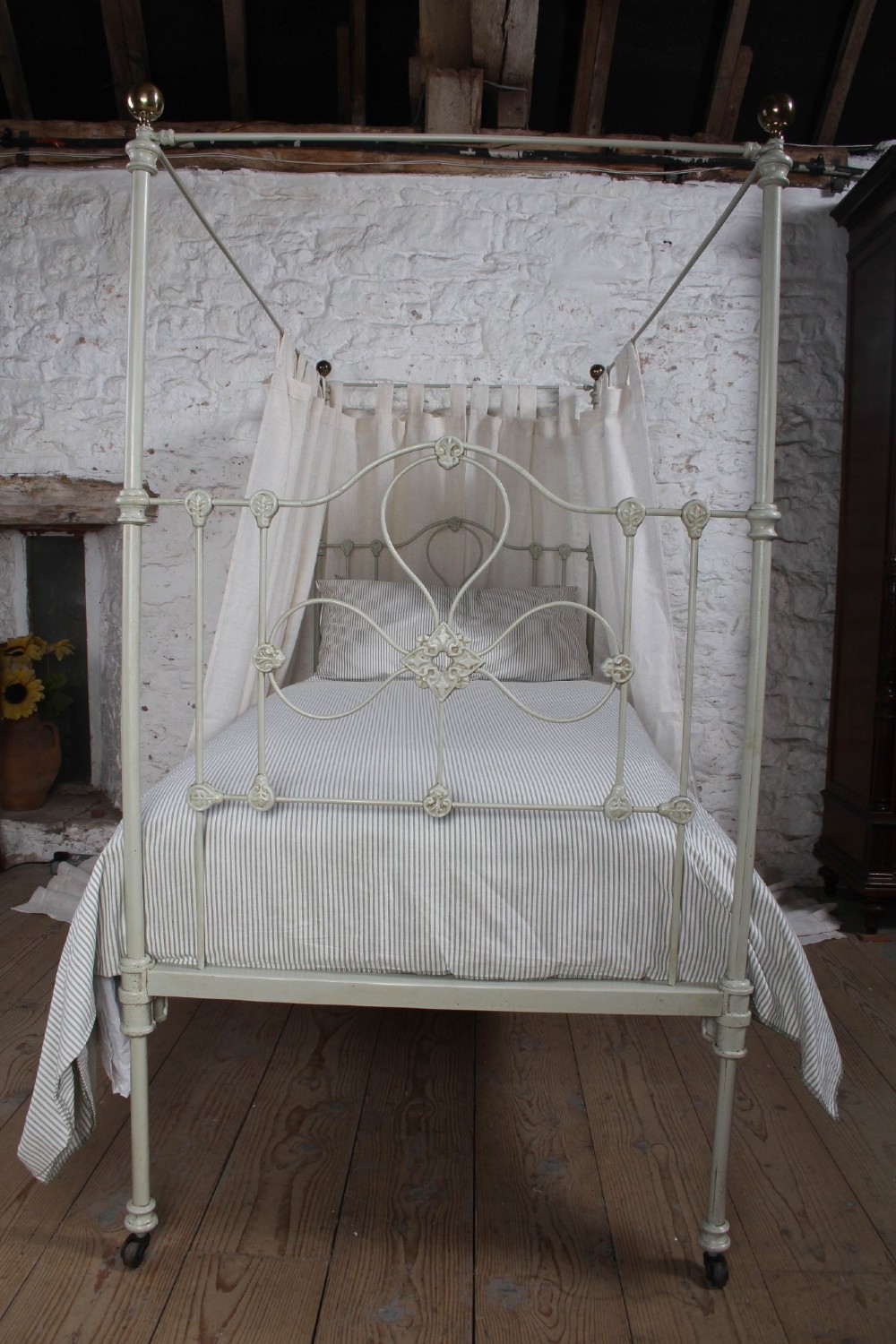 A painted single iron four-poster bed frame featuring decorative central scrollwork, brass top finials, and lightweight drapery. View VII