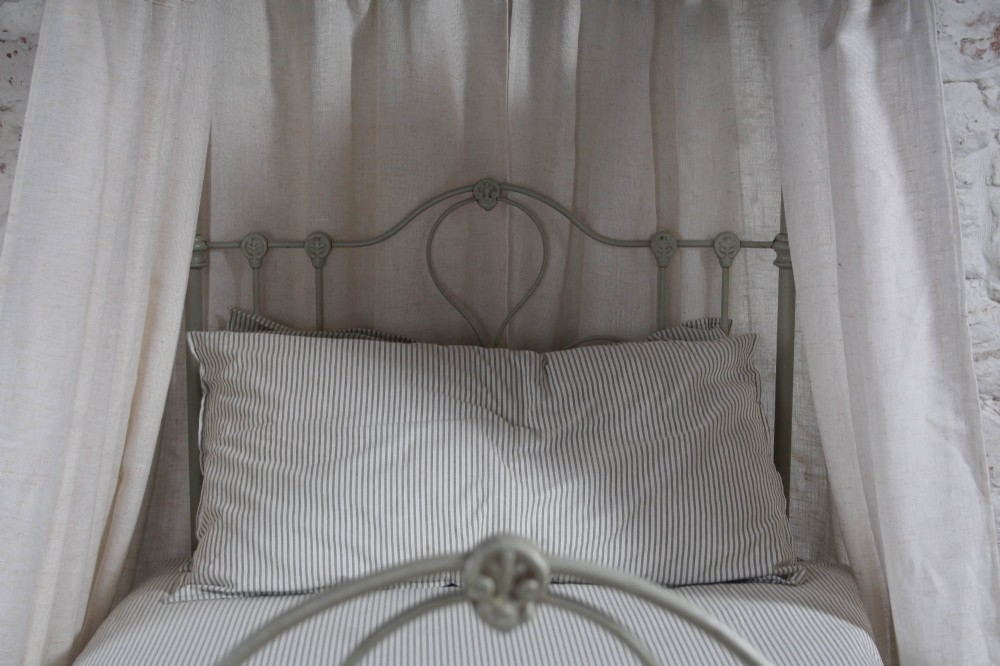 A painted single iron four-poster bed frame featuring decorative central scrollwork, brass top finials, and lightweight drapery. View VIII
