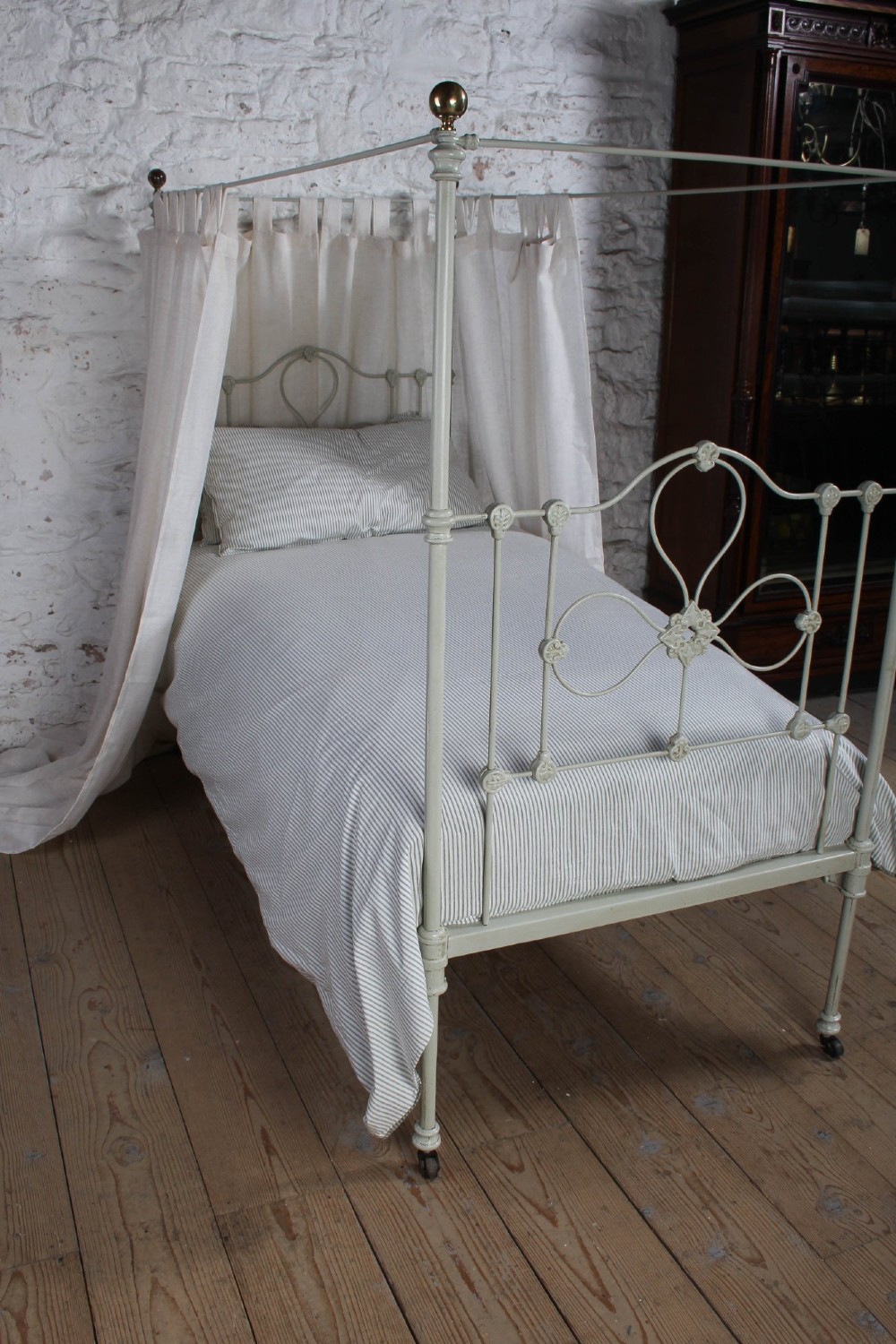 A painted single iron four-poster bed frame featuring decorative central scrollwork, brass top finials, and lightweight drapery. View IX