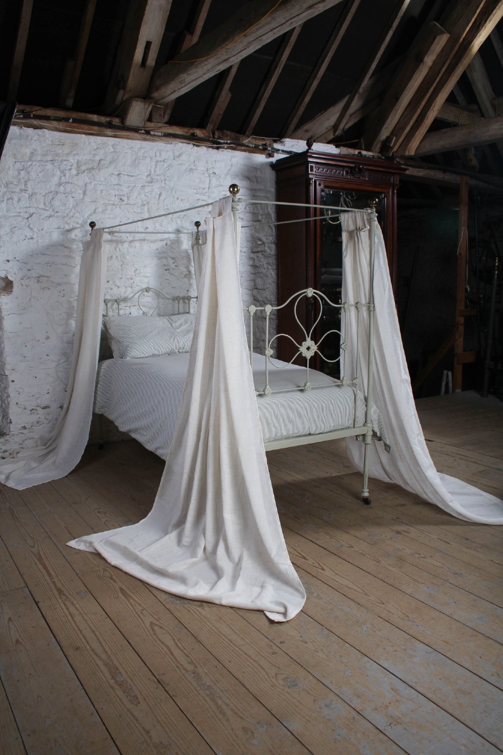 A painted single iron four-poster bed frame featuring decorative central scrollwork, brass top finials, and lightweight drapery. View XI