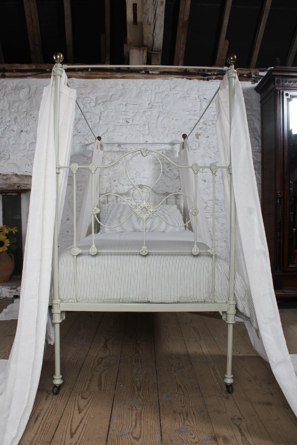 A painted single iron four-poster bed frame featuring decorative central scrollwork, brass top finials, and lightweight drapery. View XIV