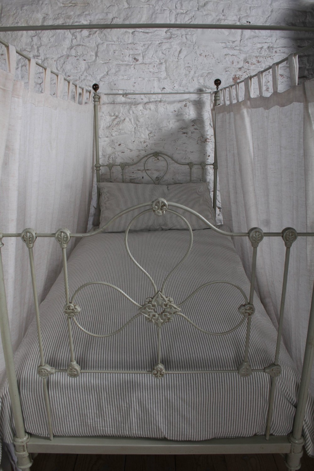 A painted single iron four-poster bed frame featuring decorative central scrollwork, brass top finials, and lightweight drapery. View XV