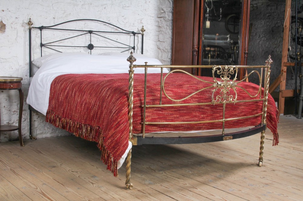 A Victorian iron and brass king-size bedstead featuring barley twist posts, ornate brass scrollwork on the footboard, and a arched black iron headboard.