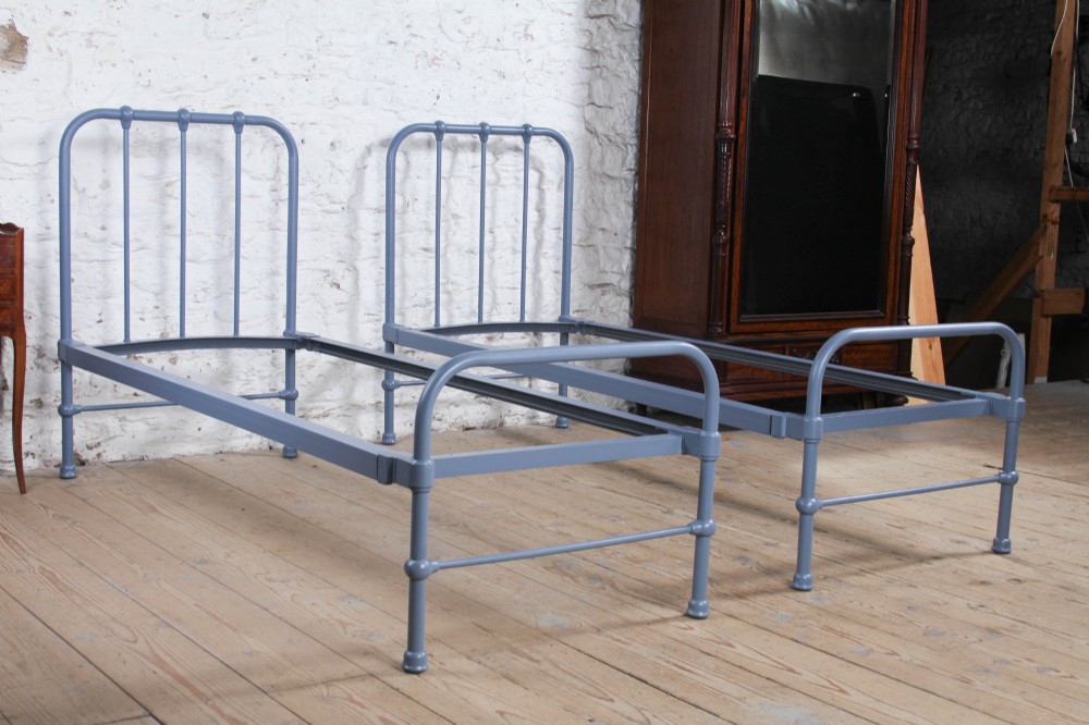 A pair of slate blue painted tubular iron single bed frames positioned side by side on a light wooden floor.