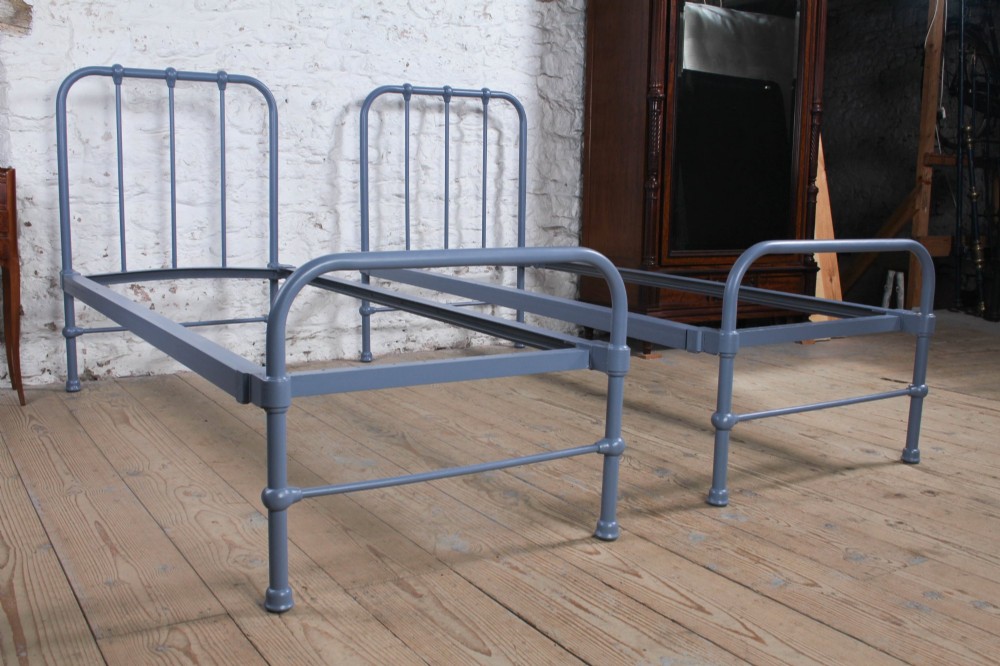 A pair of slate blue painted tubular iron single bed frames positioned side by side on a light wooden floor. View II