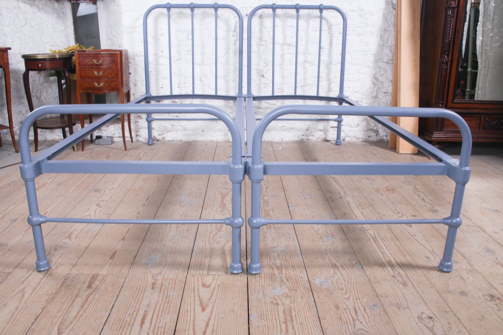 A pair of slate blue painted tubular iron single bed frames positioned side by side on a light wooden floor. View III