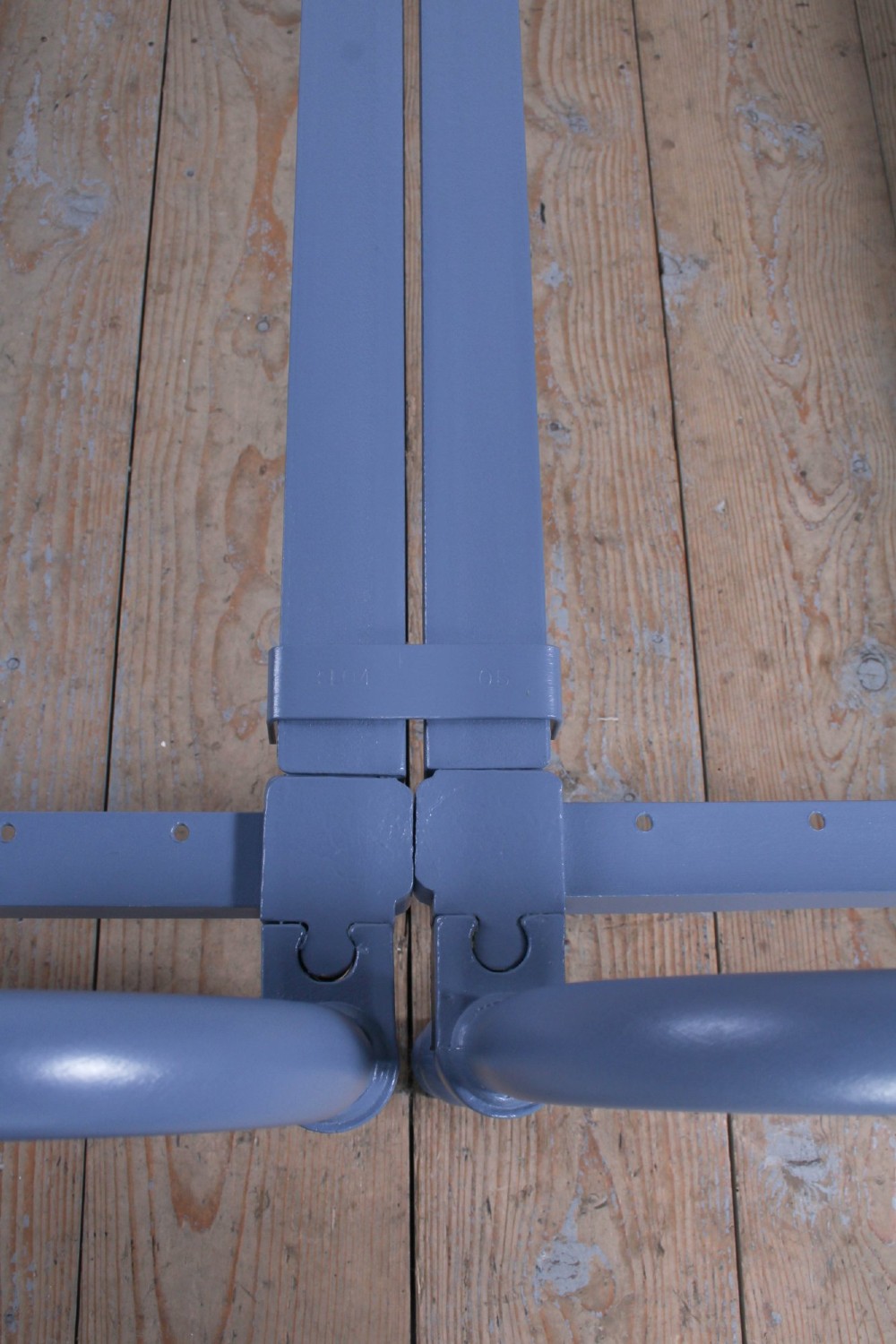 A pair of slate blue painted tubular iron single bed frames positioned side by side on a light wooden floor. View IV