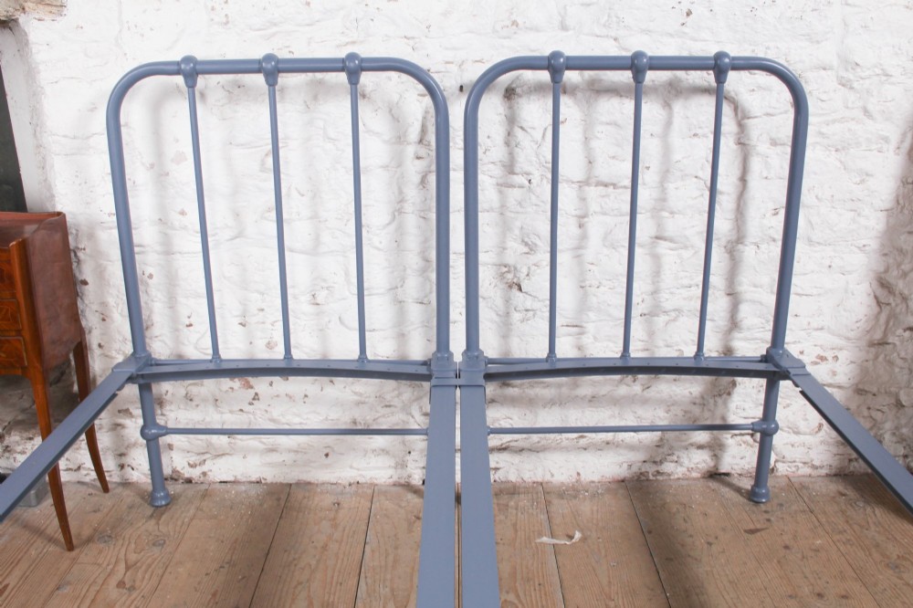 A pair of slate blue painted tubular iron single bed frames positioned side by side on a light wooden floor. View VI