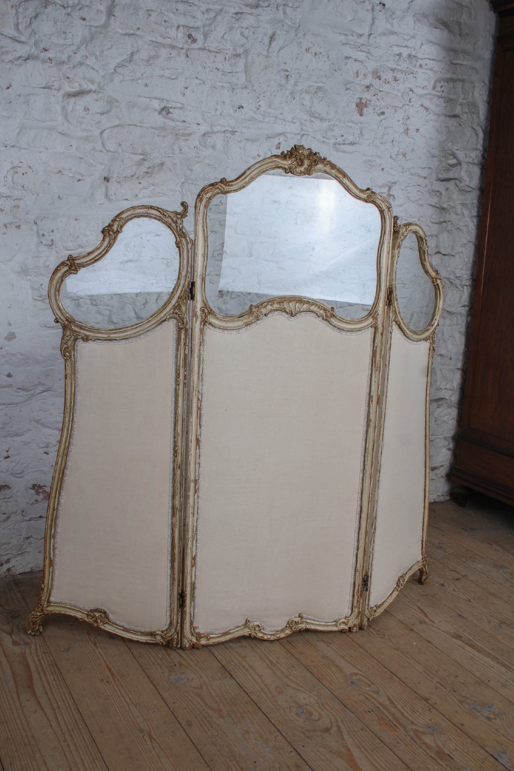 A three-panel Louis XV style folding dressing screen featuring carved cream painted timber frames with glass upper apertures and upholstered lower panels. View II