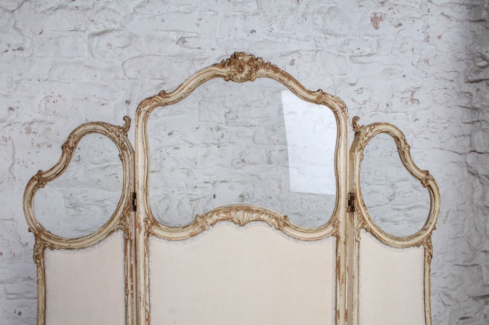 A three-panel Louis XV style folding dressing screen featuring carved cream painted timber frames with glass upper apertures and upholstered lower panels. View VI