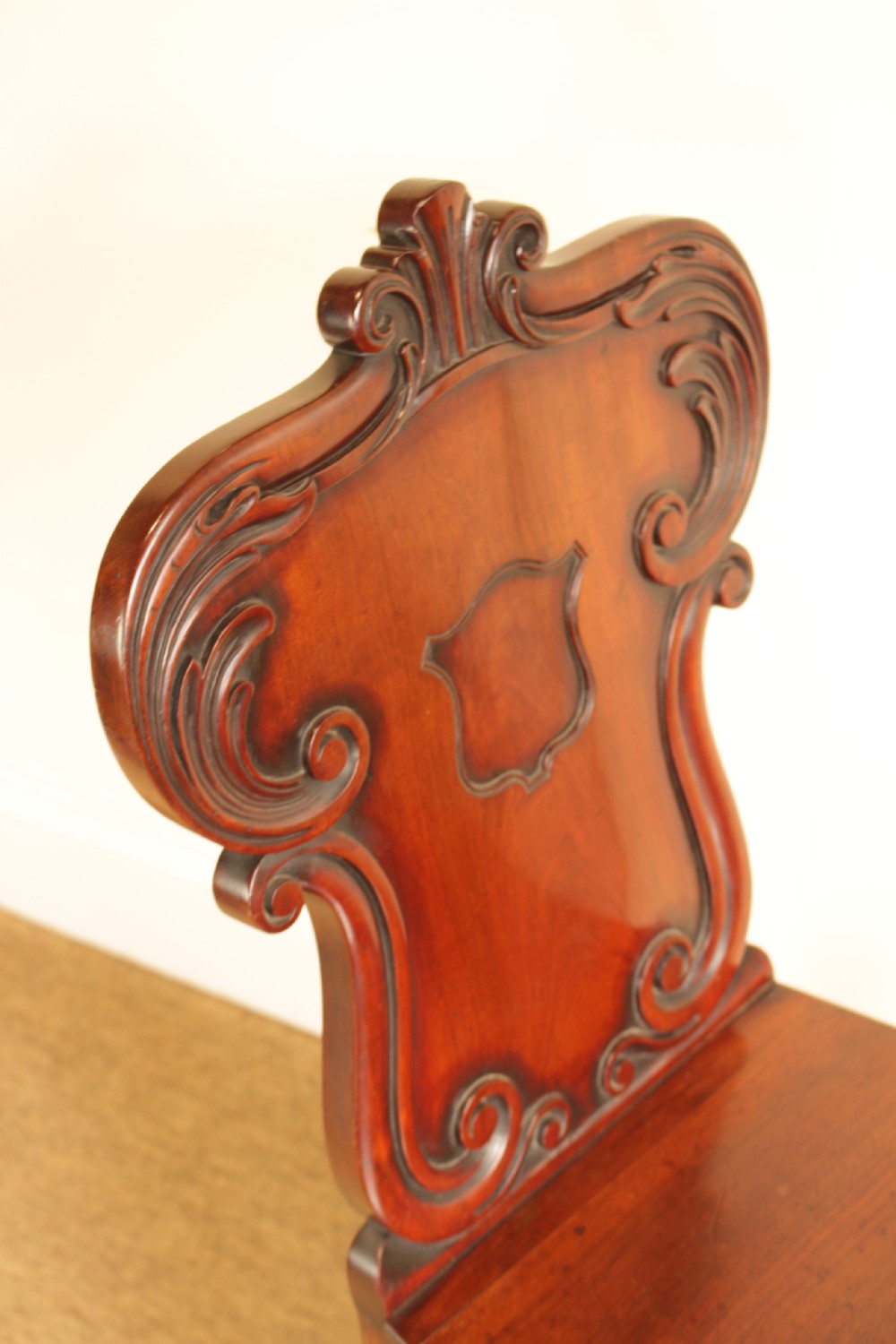 A 19th-century mahogany hall chair featuring a carved scrollwork backrest with a central shield motif, a flat wooden seat, and turned front legs. View III