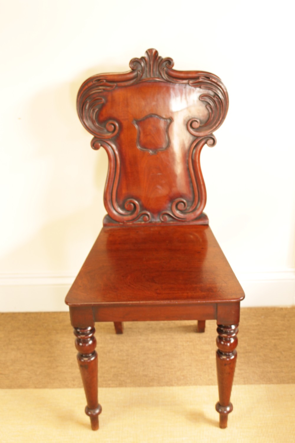 A 19th-century mahogany hall chair featuring a carved scrollwork backrest with a central shield motif, a flat wooden seat, and turned front legs. View V