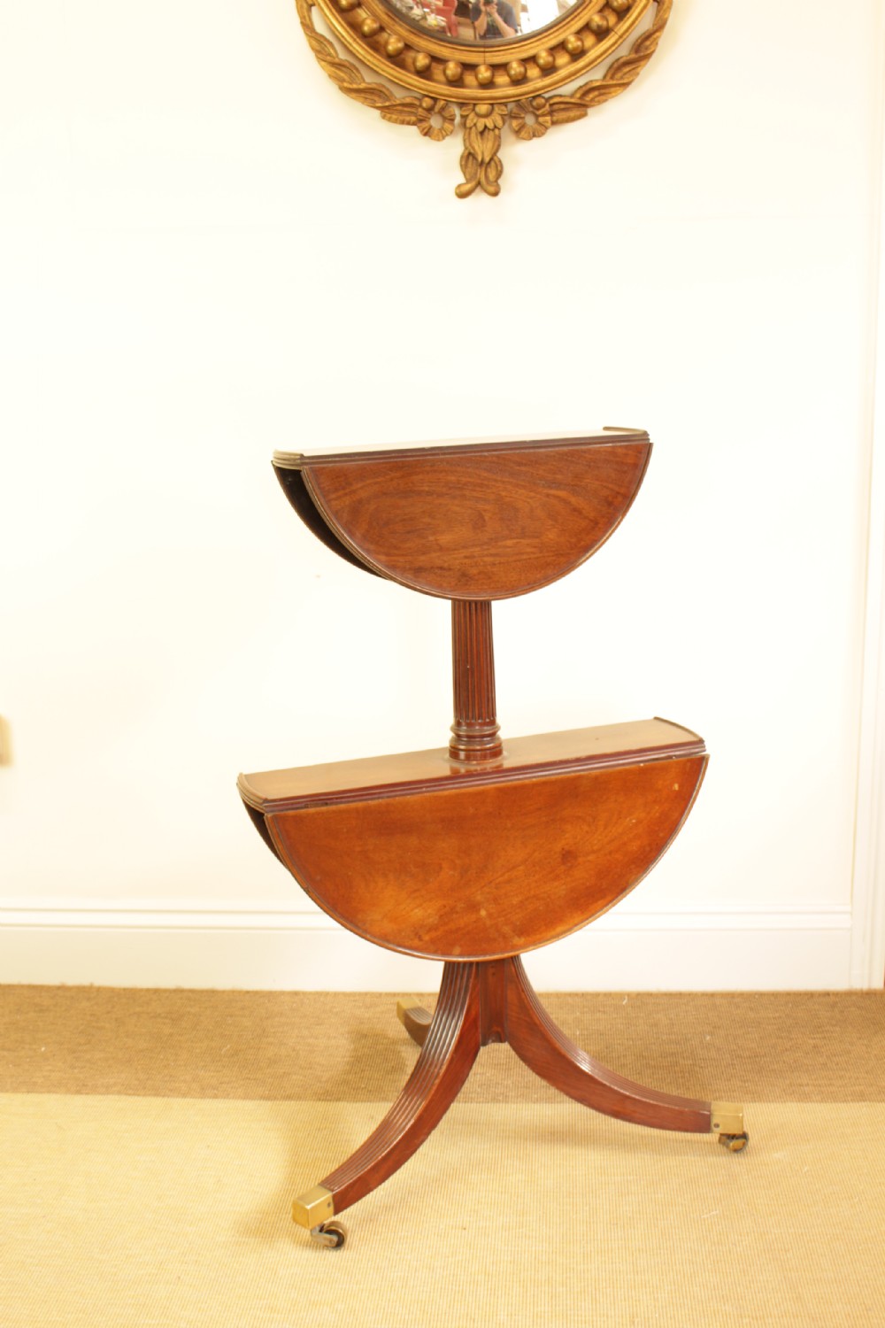 A George III mahogany two-tiered dumb waiter with folding drop-leaf sides, fluted central pillar, and tripod reeded legs on brass castors.