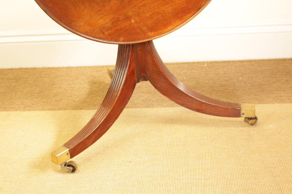 A George III mahogany two-tiered dumb waiter with folding drop-leaf sides, fluted central pillar, and tripod reeded legs on brass castors. View IV