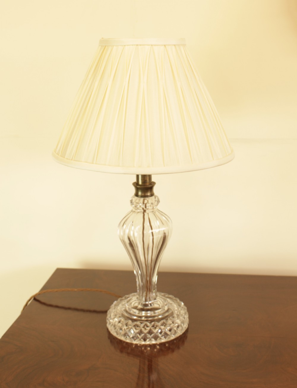 A cut crystal table lamp featuring a baluster-shaped glass base and a cream pleated fabric conical lampshade.