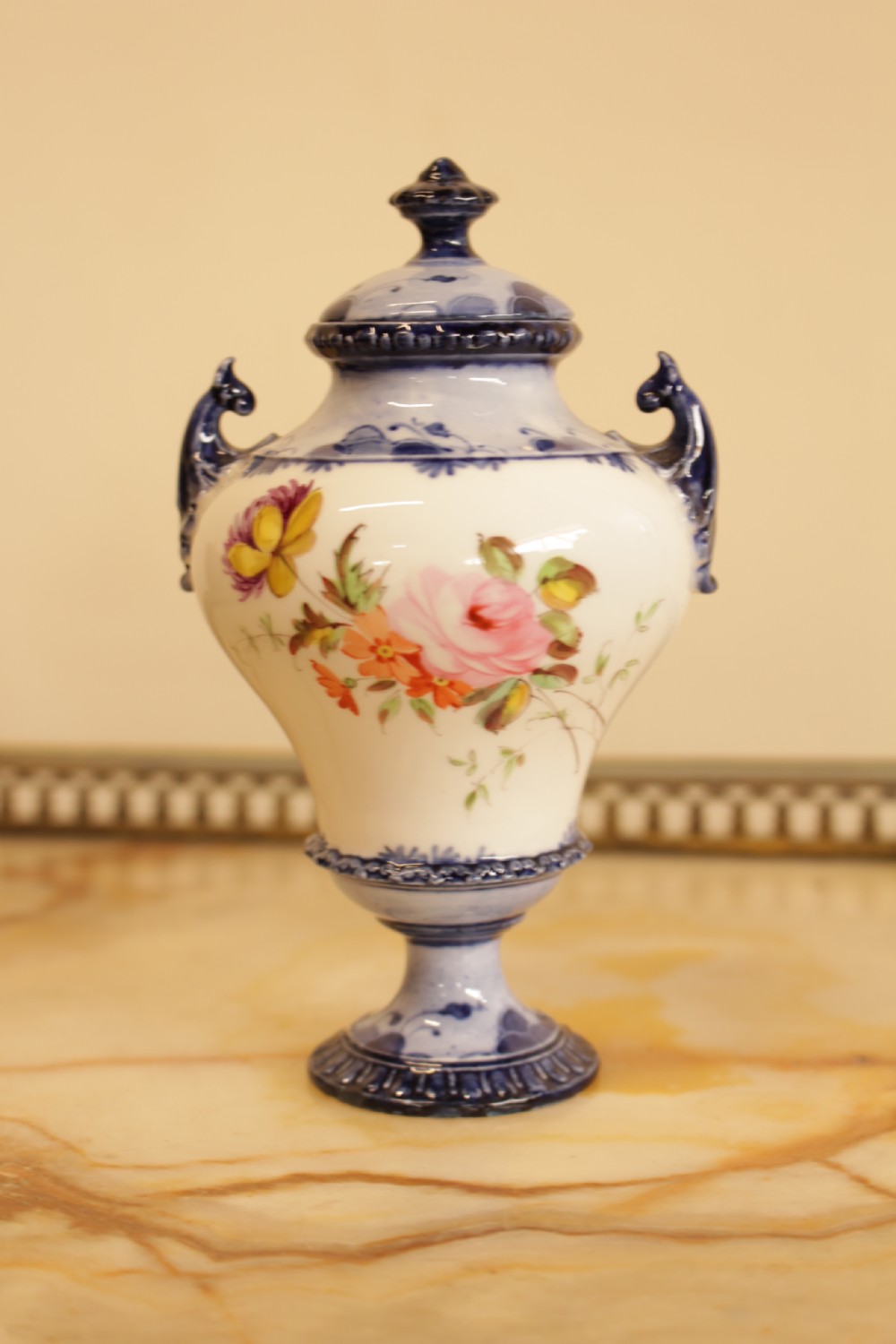 An antique porcelain lidded urn featuring hand-painted floral motifs, cobalt blue handles, and a pedestal base.