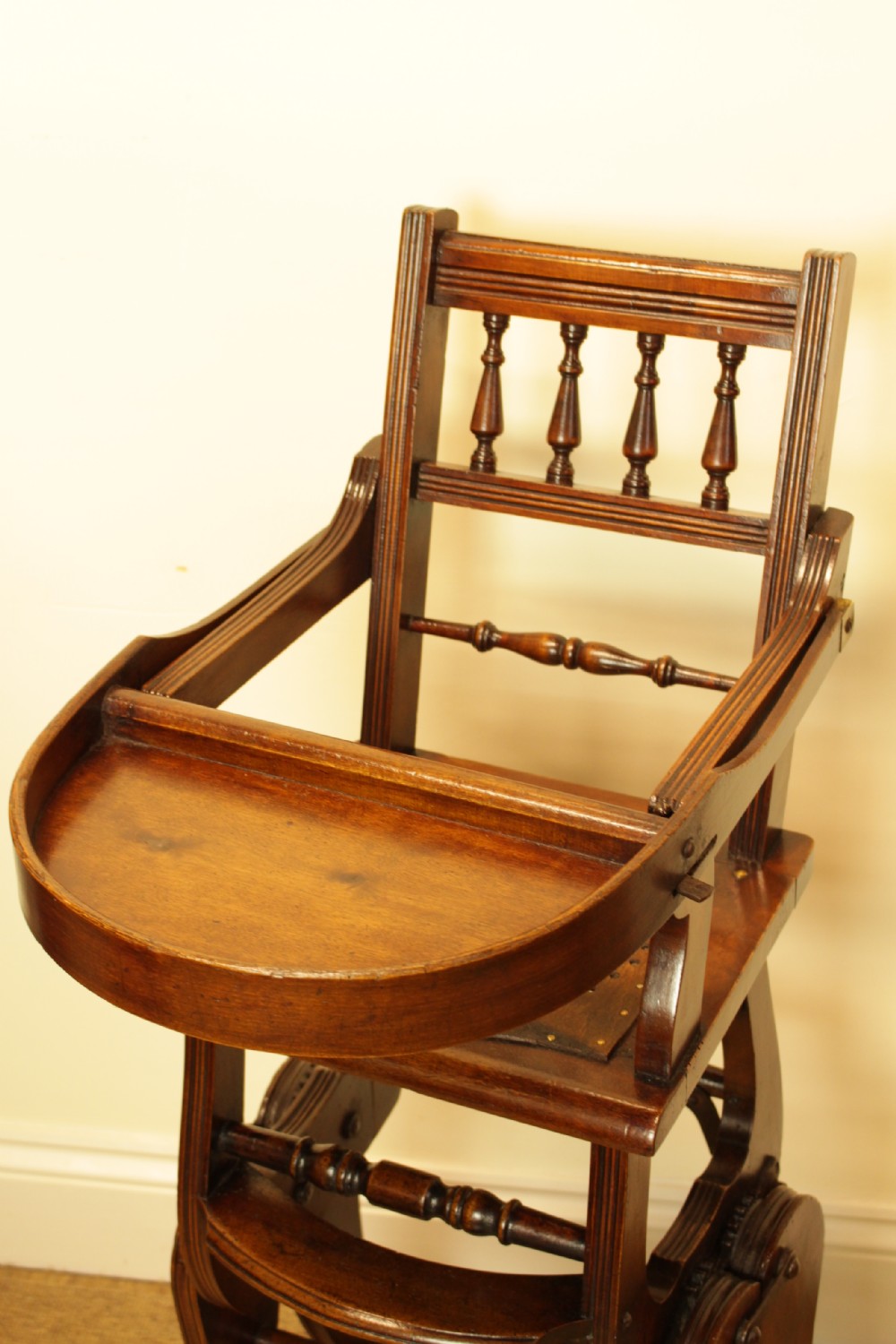 A Victorian dark brown beech metamorphic childs high chair featuring turned spindles, a perforated seat, and small wooden wheels. View II