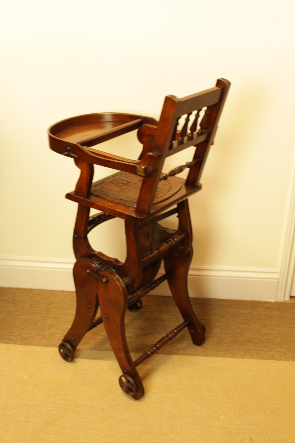 A Victorian dark brown beech metamorphic childs high chair featuring turned spindles, a perforated seat, and small wooden wheels. View III