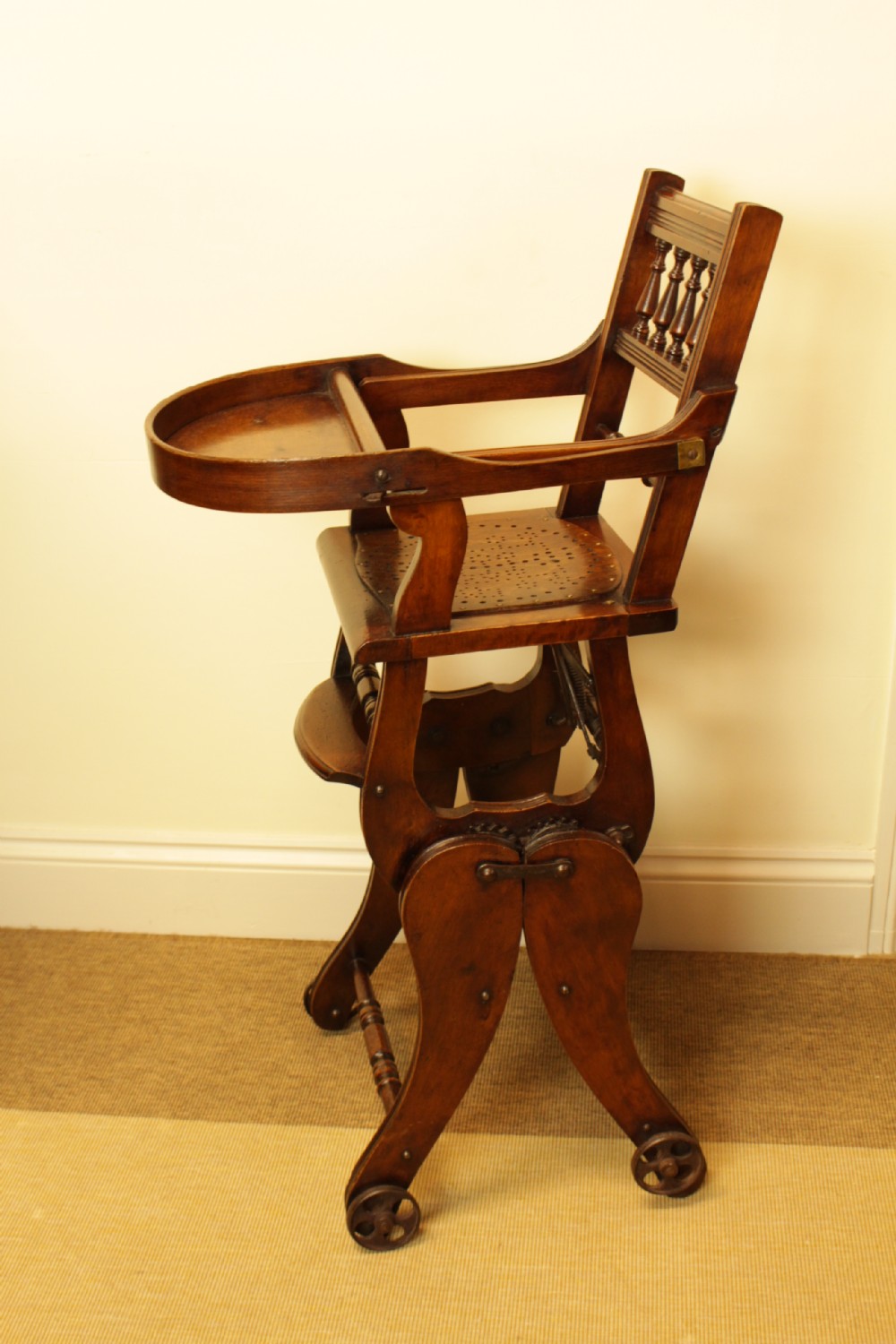 A Victorian dark brown beech metamorphic childs high chair featuring turned spindles, a perforated seat, and small wooden wheels. View V