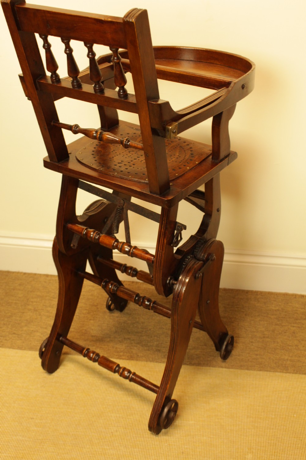 A Victorian dark brown beech metamorphic childs high chair featuring turned spindles, a perforated seat, and small wooden wheels. View VII
