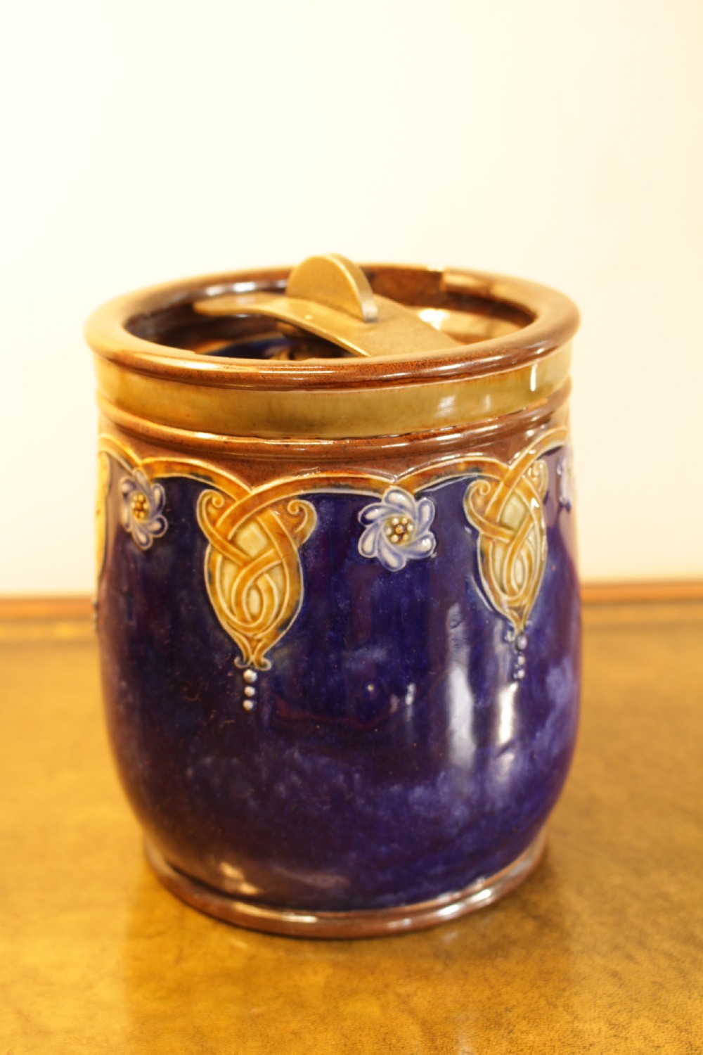 A cylindrical Royal Doulton stoneware tobacco jar decorated with a deep blue glaze, ochre stylized floral motifs, and a metal press-lid mechanism.
