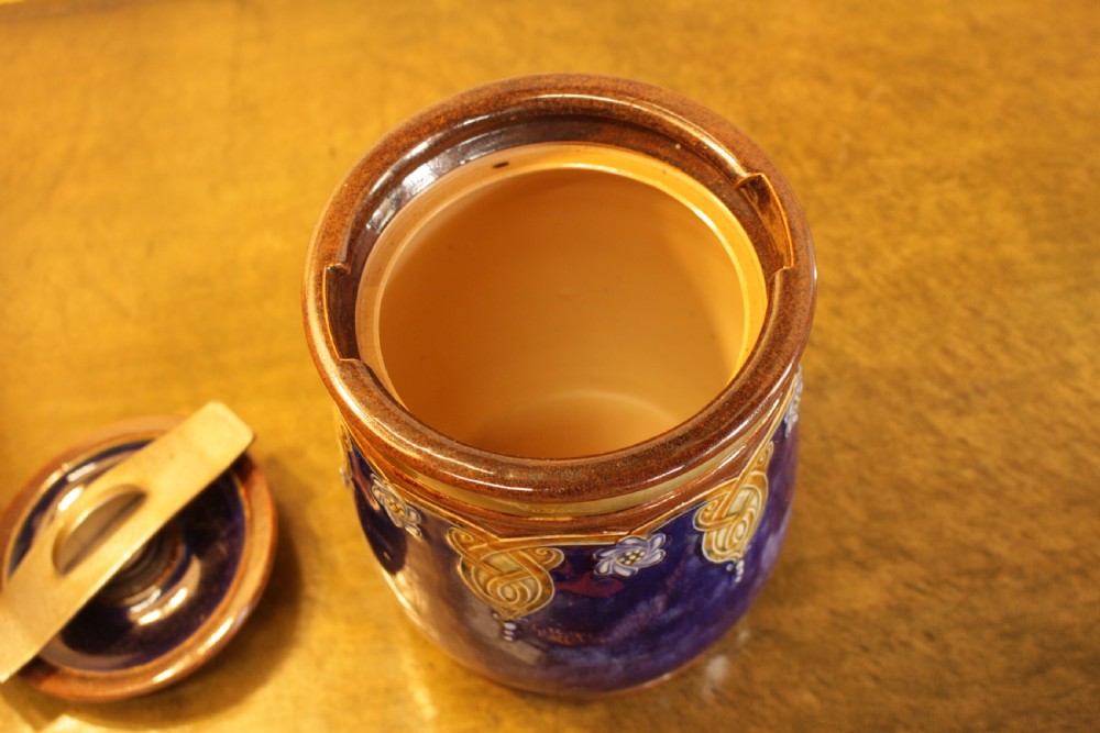 A cylindrical Royal Doulton stoneware tobacco jar decorated with a deep blue glaze, ochre stylized floral motifs, and a metal press-lid mechanism. View III