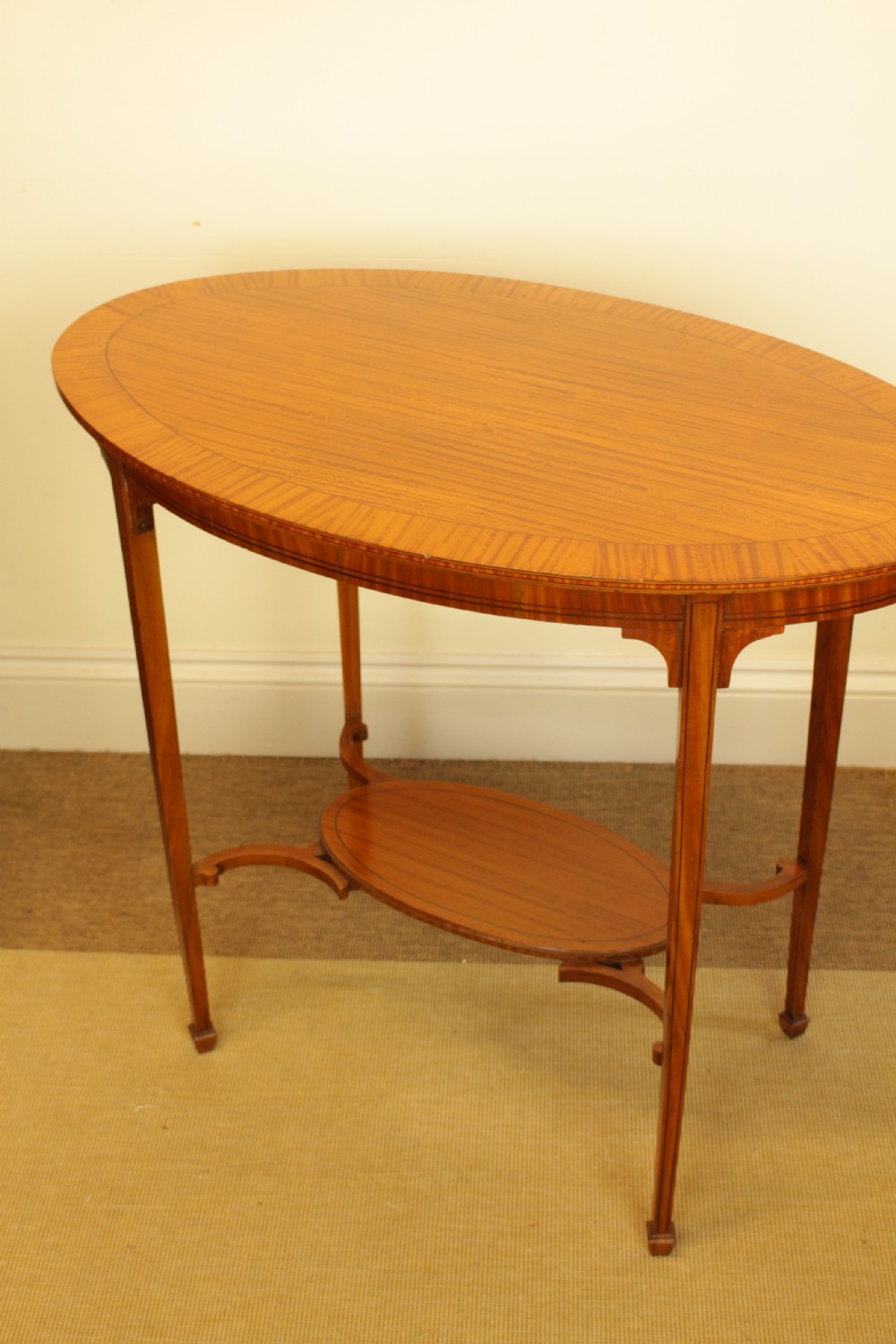 An antique oval satinwood occasional table featuring a crossbanded veneered top, slender tapered legs, and a matching lower undertier shelf. View III