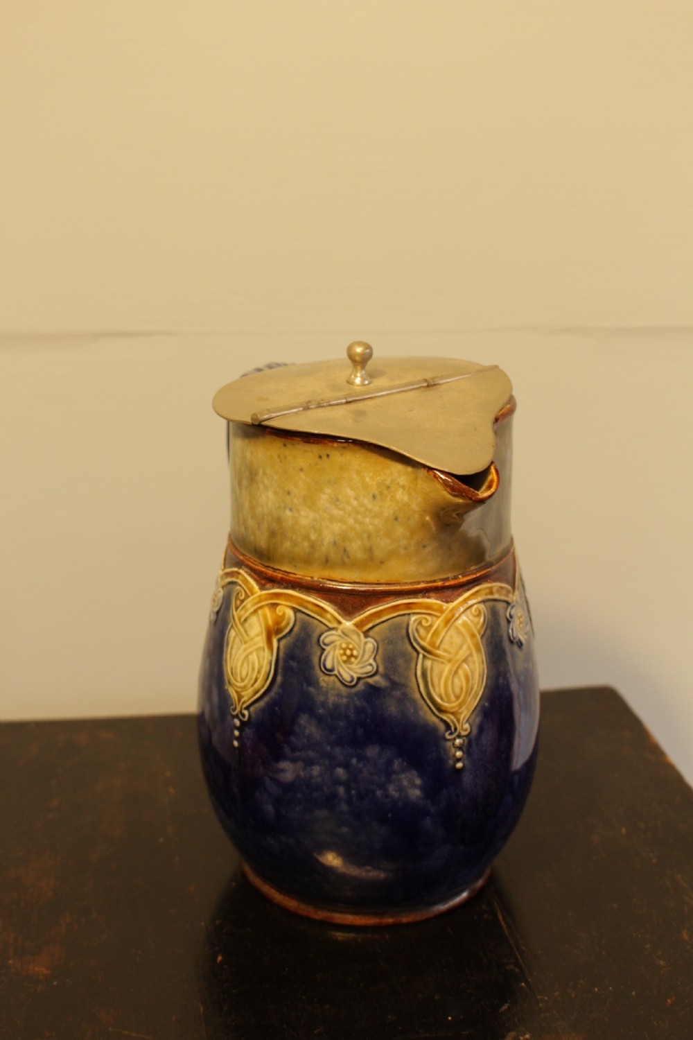A Royal Doulton salt-glazed stoneware water jug featuring a cobalt blue body with raised relief scrollwork, an ochre neck, and a hinged metal lid. View III