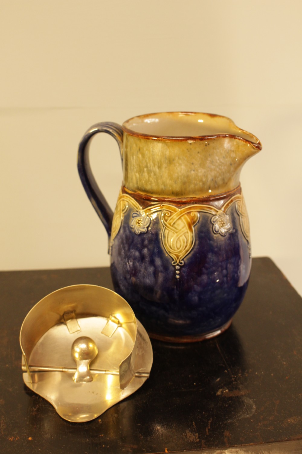 A Royal Doulton salt-glazed stoneware water jug featuring a cobalt blue body with raised relief scrollwork, an ochre neck, and a hinged metal lid. View VI