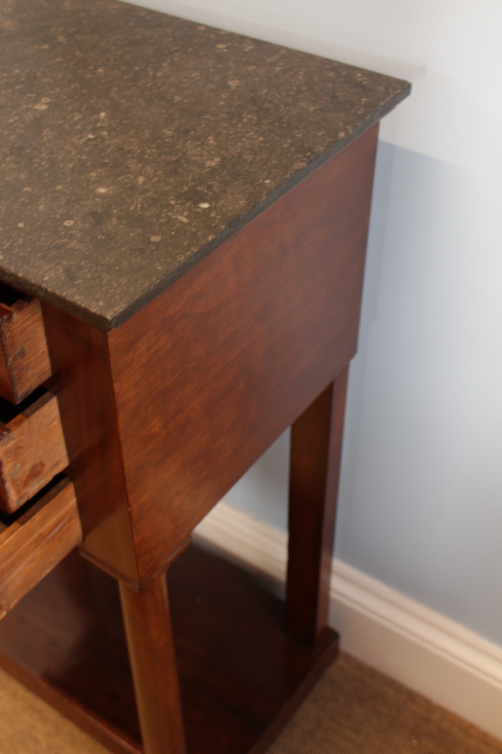 A French Empire flame mahogany side table featuring a dark grey marble top, three drawers with brass keyholes, turned column front legs, and an undertier shelf. View II