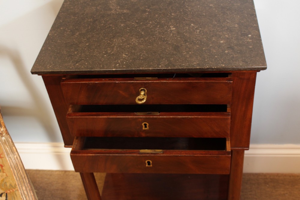 A French Empire flame mahogany side table featuring a dark grey marble top, three drawers with brass keyholes, turned column front legs, and an undertier shelf. View V