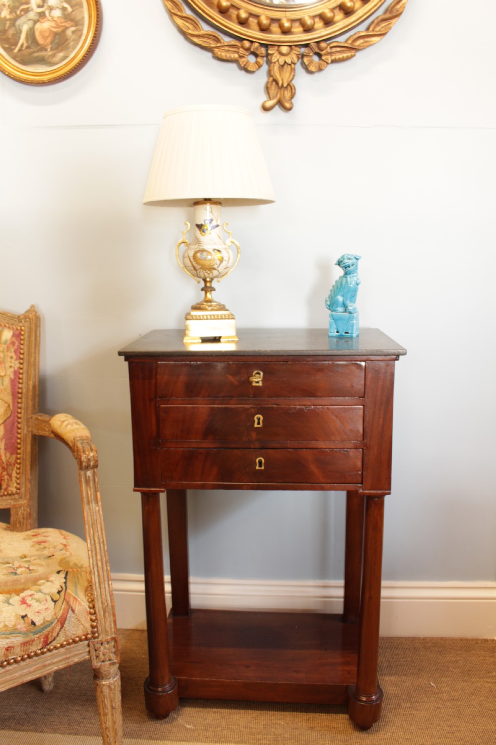 A French Empire flame mahogany side table featuring a dark grey marble top, three drawers with brass keyholes, turned column front legs, and an undertier shelf. View IX