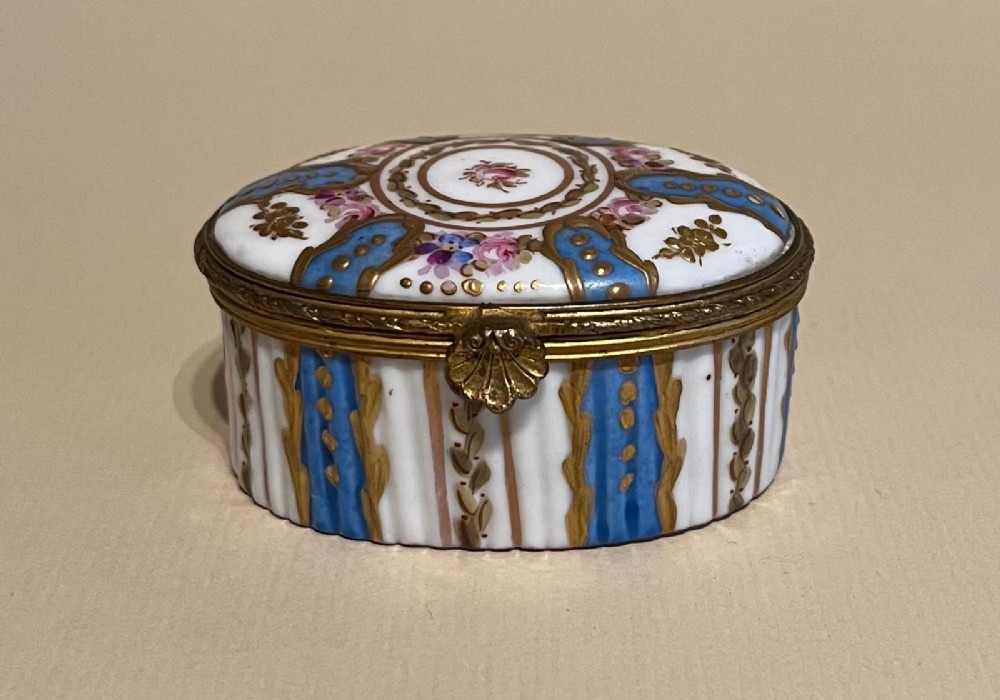 An oval late nineteenth-century hand-painted white porcelain trinket box featuring blue stripes, floral motifs, gilt decoration, and a brass shell clasp.