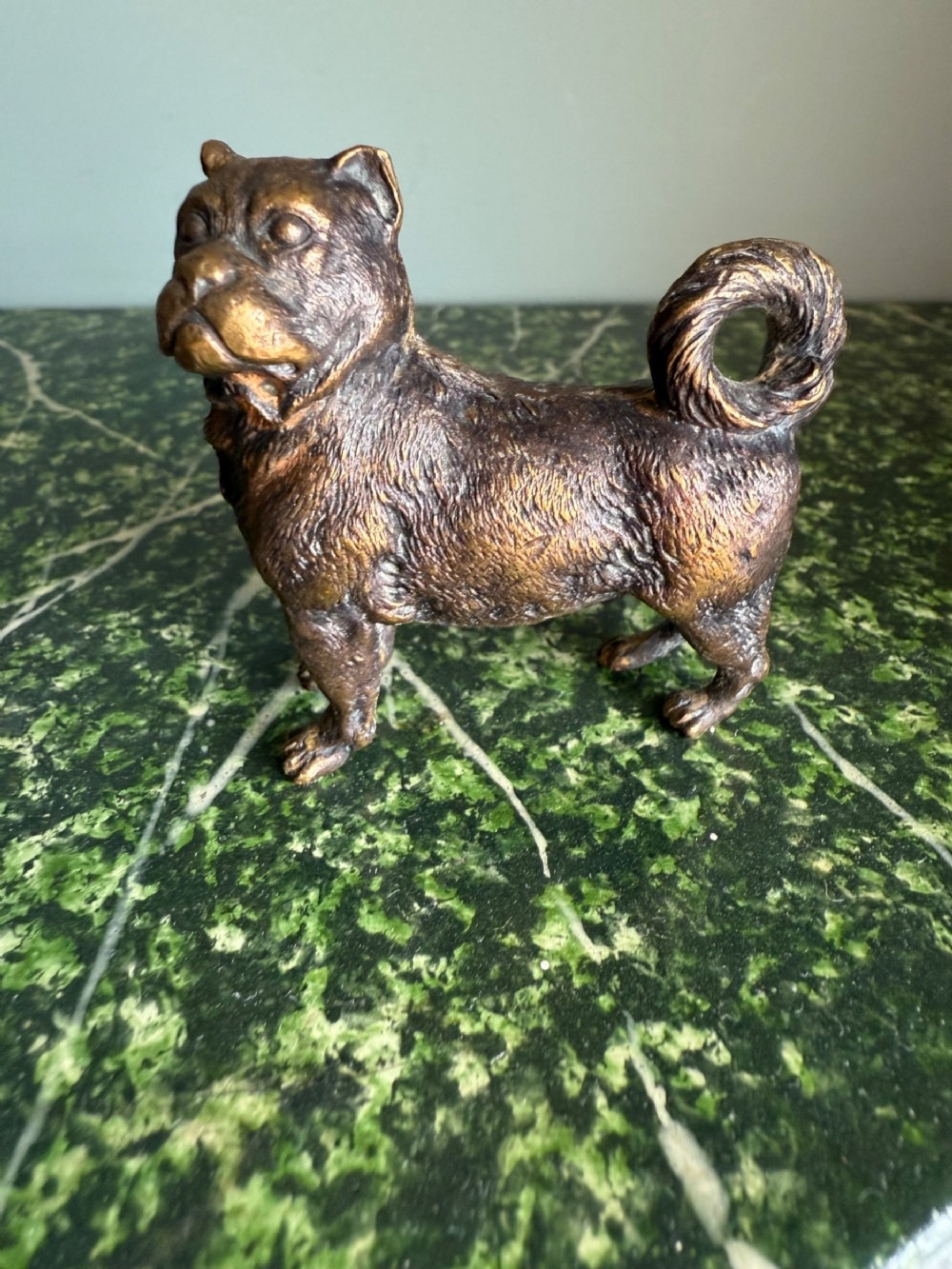 A small bronze figurine depicting a standing pug dog with a textured coat and curled tail.