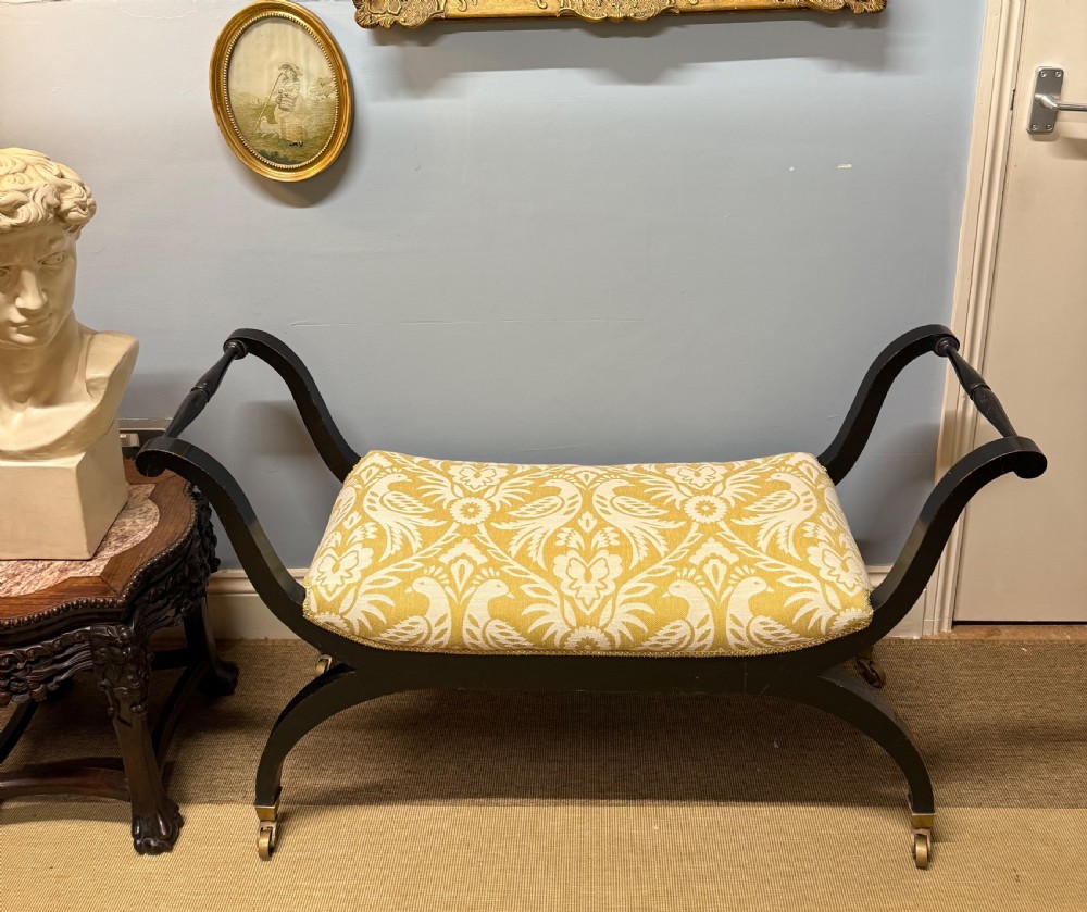 An Edwardian ebonised wooden window seat featuring curved swept arms, brass castors, and a patterned yellow upholstered cushion.
