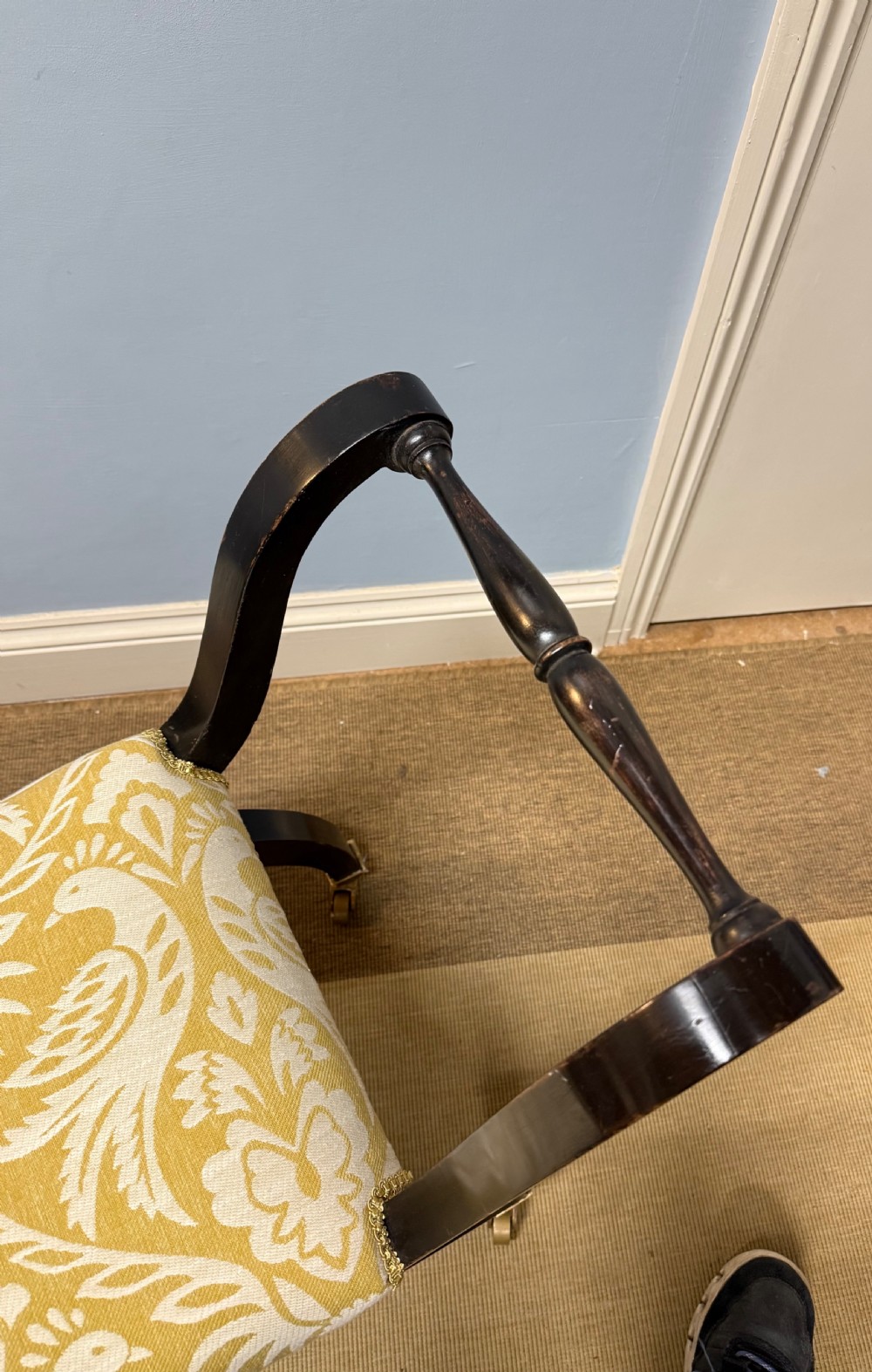 An Edwardian ebonised wooden window seat featuring curved swept arms, brass castors, and a patterned yellow upholstered cushion. View II