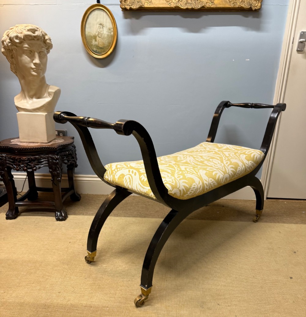 An Edwardian ebonised wooden window seat featuring curved swept arms, brass castors, and a patterned yellow upholstered cushion. View IV