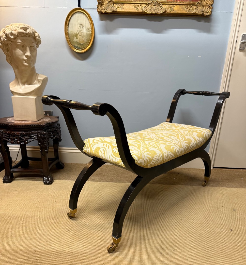 An Edwardian ebonised wooden window seat featuring curved swept arms, brass castors, and a patterned yellow upholstered cushion. View V