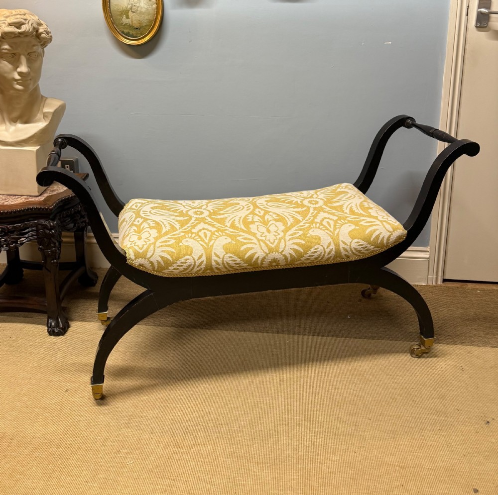 An Edwardian ebonised wooden window seat featuring curved swept arms, brass castors, and a patterned yellow upholstered cushion. View VII