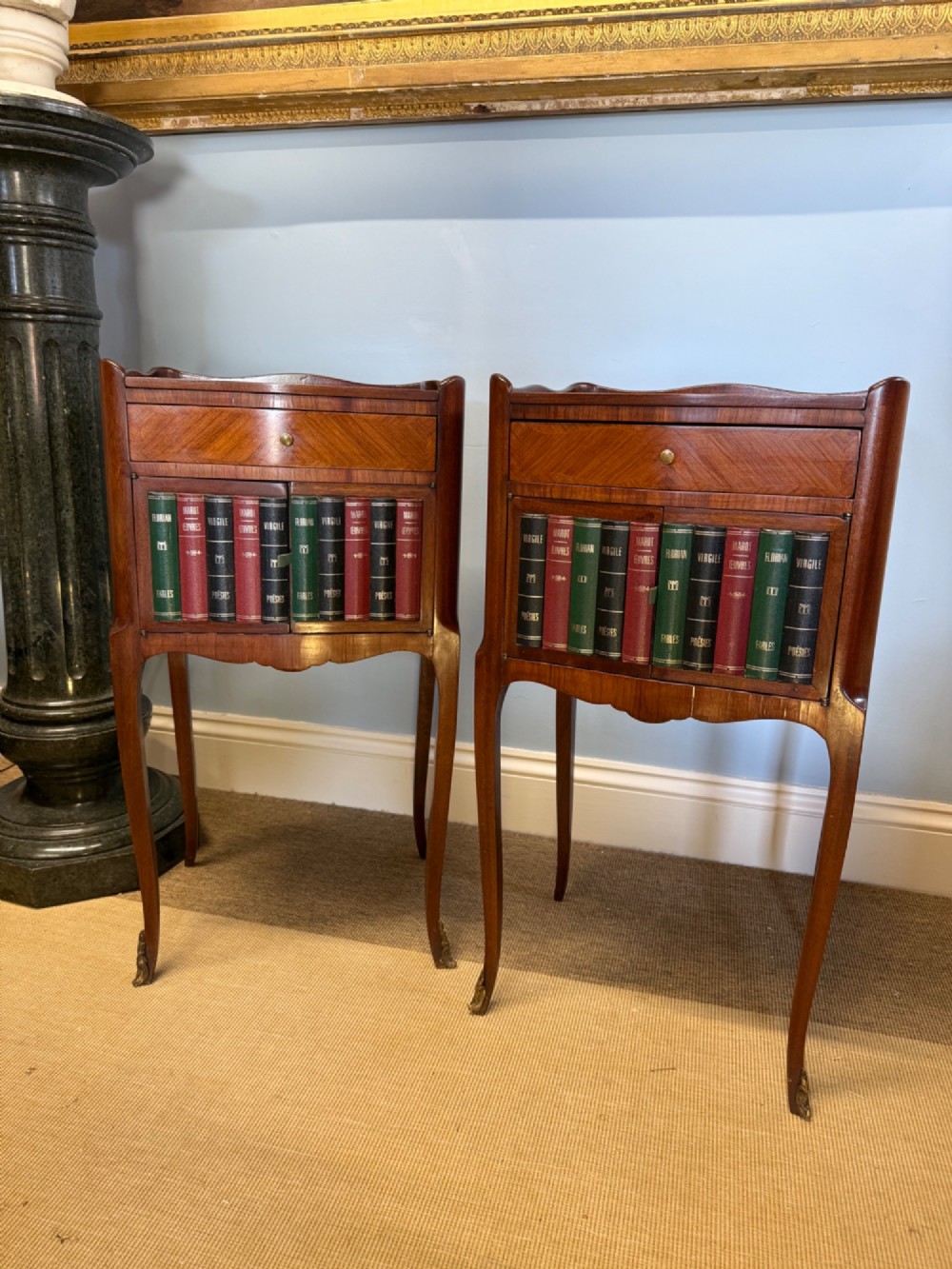 A pair of French walnut bedside tables featuring faux book spine cabinet doors, single drawers with chevron veneer patterns, and slender cabriole legs ending in brass sabots.