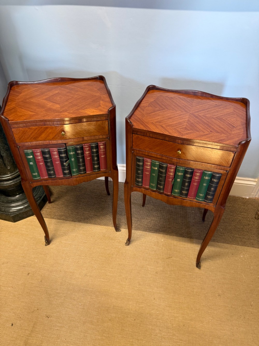 A pair of French walnut bedside tables featuring faux book spine cabinet doors, single drawers with chevron veneer patterns, and slender cabriole legs ending in brass sabots. View II
