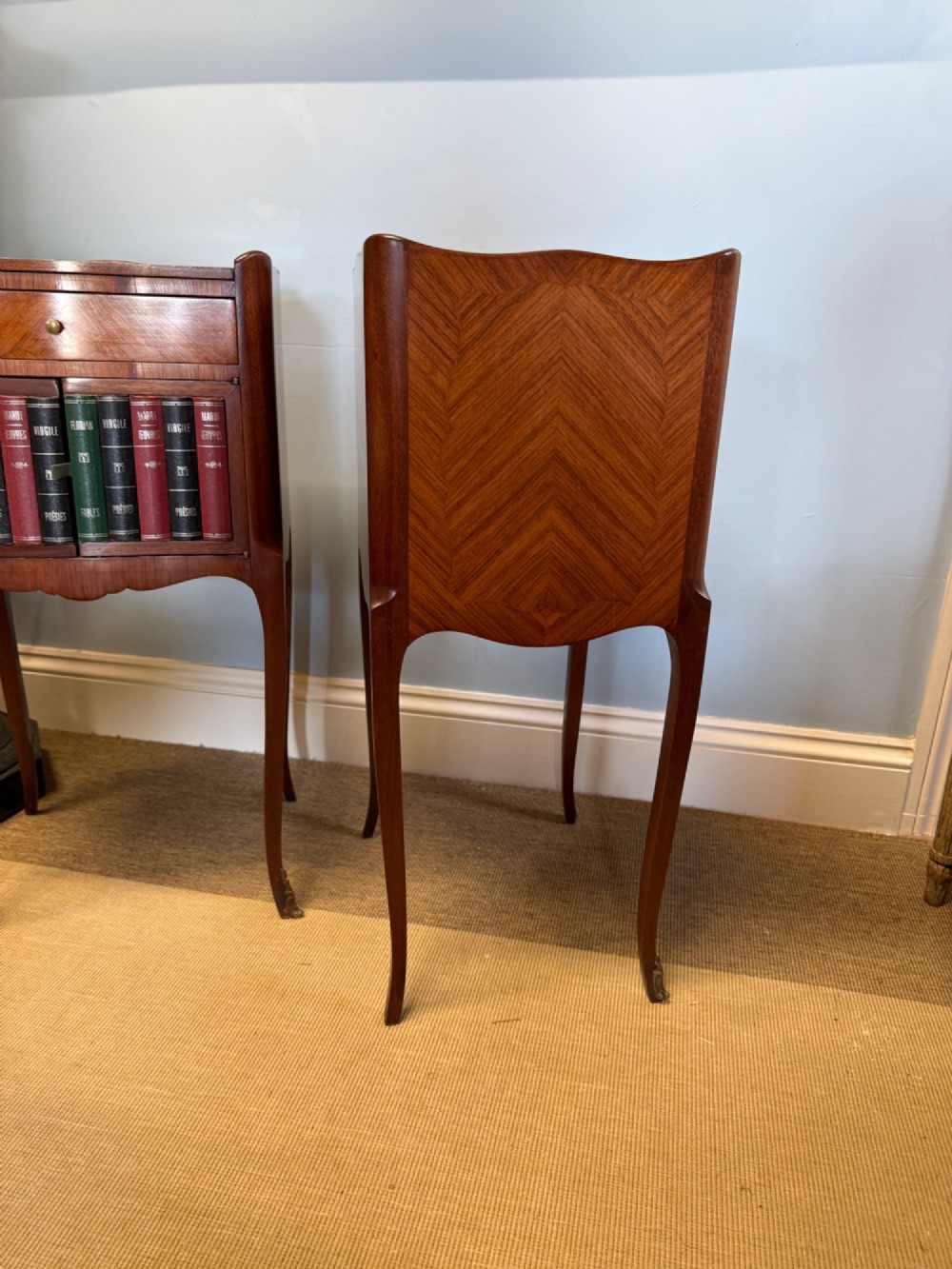 A pair of French walnut bedside tables featuring faux book spine cabinet doors, single drawers with chevron veneer patterns, and slender cabriole legs ending in brass sabots. View IV