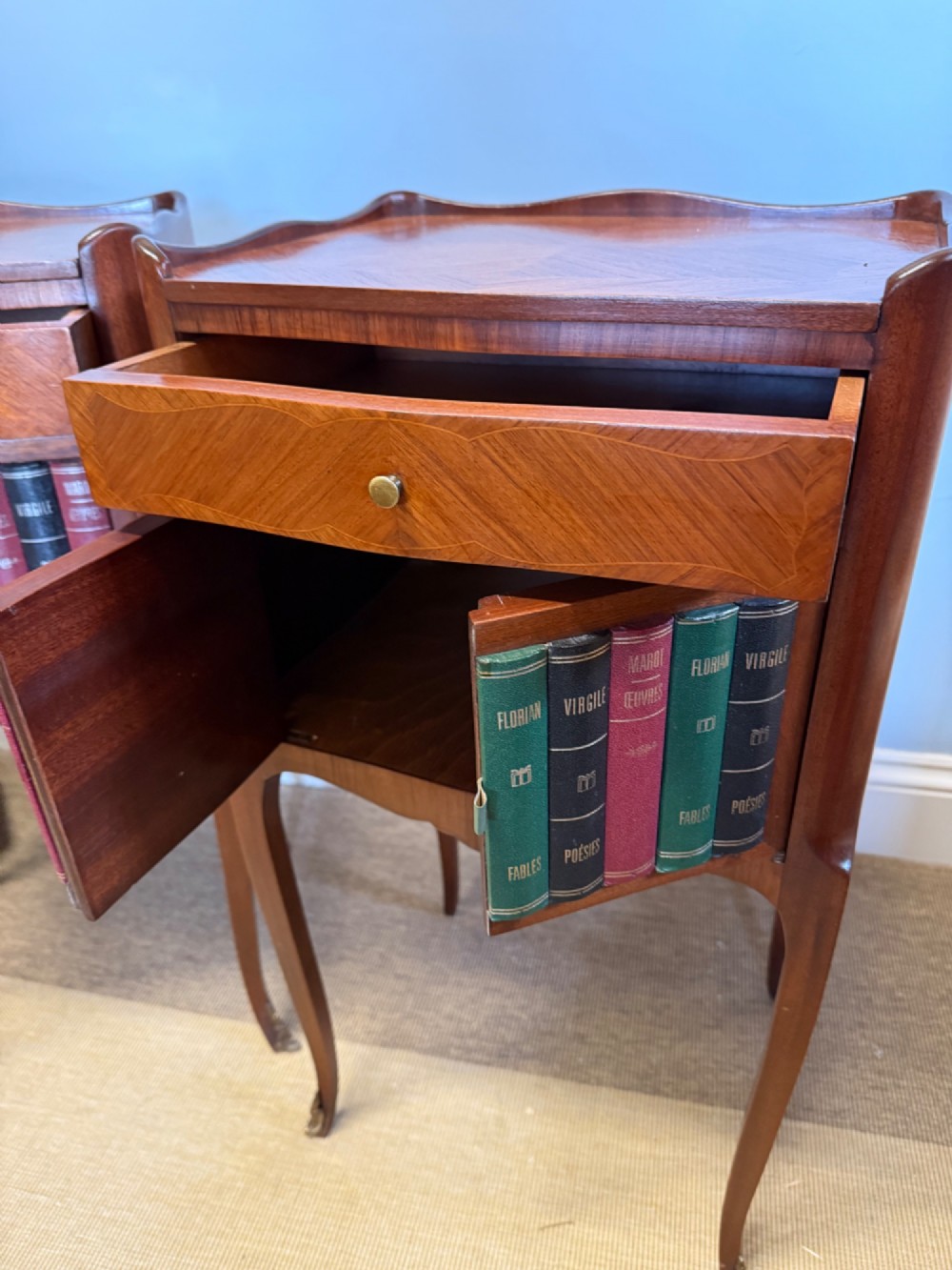 A pair of French walnut bedside tables featuring faux book spine cabinet doors, single drawers with chevron veneer patterns, and slender cabriole legs ending in brass sabots. View V