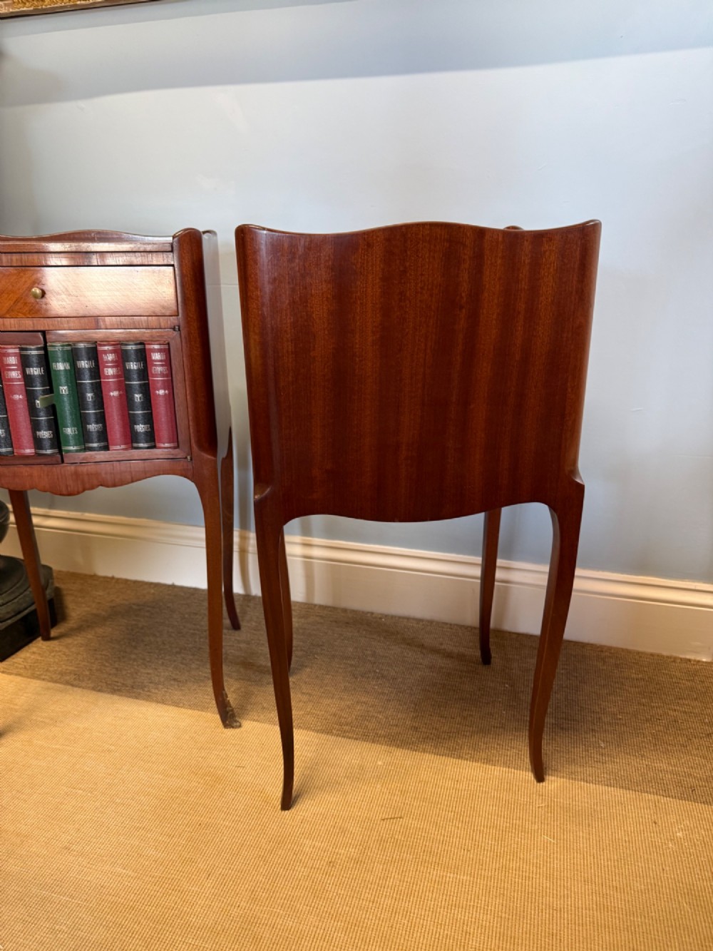 A pair of French walnut bedside tables featuring faux book spine cabinet doors, single drawers with chevron veneer patterns, and slender cabriole legs ending in brass sabots. View VI