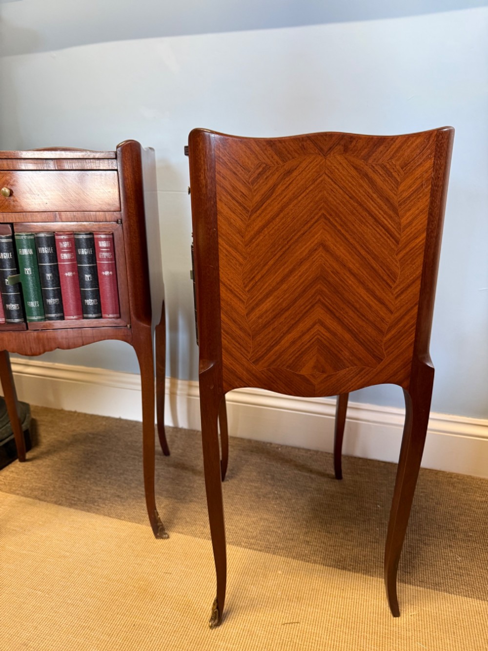 A pair of French walnut bedside tables featuring faux book spine cabinet doors, single drawers with chevron veneer patterns, and slender cabriole legs ending in brass sabots. View VII